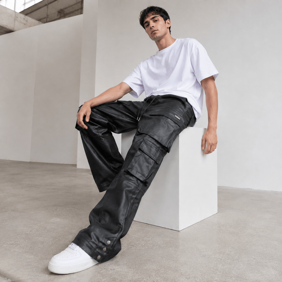 Seated model wearing black cargo pants, a white T-shirt and white sneakers in a minimal studio.
