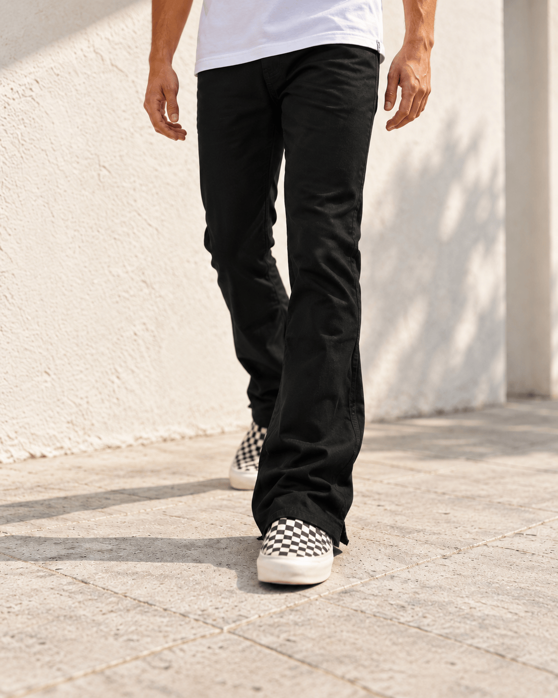 Waist-down view of a model walking in black flare pants beside a sunlit white wall.